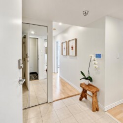 Foyer of an apartment with a 1205 plaque, mirrored wall, wooden bench, and a white orchid plant on a small table edge.