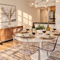 Round marble dining table set for six with clear acrylic chairs, wine bottles, candles, and a succulent centerpiece in a sunny modern dining area