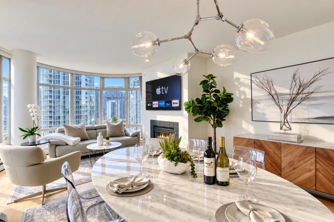 Modern open-concept living and dining area with a marble table set for four, wine bottles, plants, and a fireplace under a chandelier.