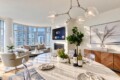 Modern open-concept living and dining area with a marble table set for four, wine bottles, plants, and a fireplace under a chandelier.