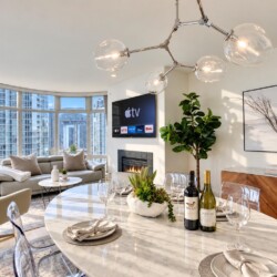 Modern open-concept living and dining area with a marble table set for four, wine bottles, plants, and a fireplace under a chandelier.