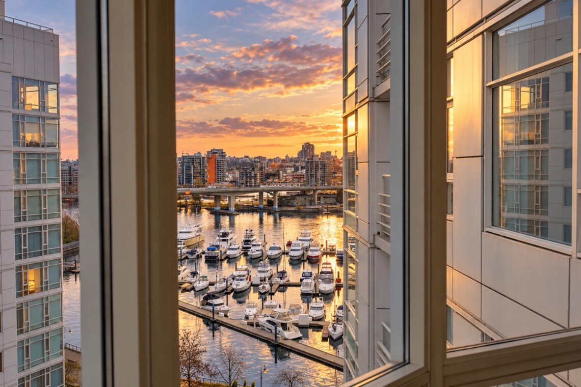 Sunset over a marina viewed from a window, with boats docked and a city skyline in the distance.