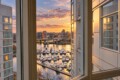 Sunset over a marina viewed from a window, with boats docked and a city skyline in the distance.