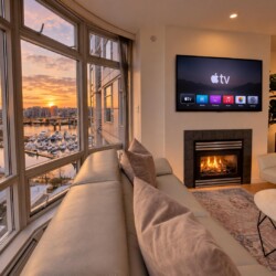 Living room with oversized curved windows showing a marina and sunset, beige sofa, and a fireplace with a wall-mounted TV above it