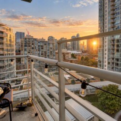 View from a high-rise balcony overlooking a cityscape at sunset, with string lights along the railing in the foreground.