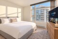 Bright hotel bedroom with a king bed, white duvet and beige pillows, sunlit from a large window, and a flat-screen TV on a wooden dresser nearby.