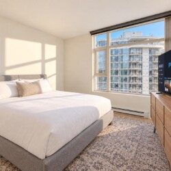 Bright hotel bedroom with a king bed, white duvet and beige pillows, sunlit from a large window, and a flat-screen TV on a wooden dresser nearby.