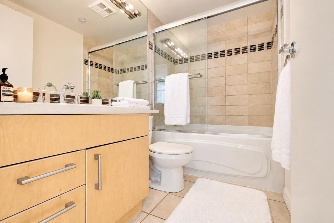 Modern bathroom with a light wood vanity, white countertop, and chrome fixtures; glass shower doors and beige tiled walls.