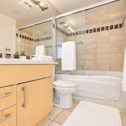 Modern bathroom with a light wood vanity, white countertop, and chrome fixtures; glass shower doors and beige tiled walls.