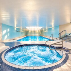 Indoor spa facility with a round bubbling hot tub in the foreground and a rectangular pool beyond.