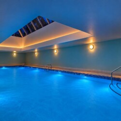 Indoor pool area with bright blue water, blue walls, and a skylight in the ceiling; warm wall and ceiling lighting creates a spa-like ambiance.