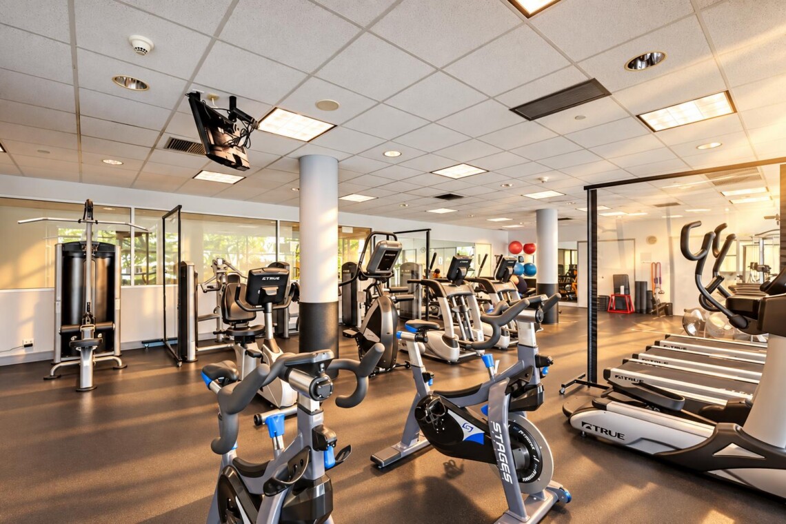 Modern gym with cardio machines (stationary bikes and treadmills), large windows, and mirrors in the background.