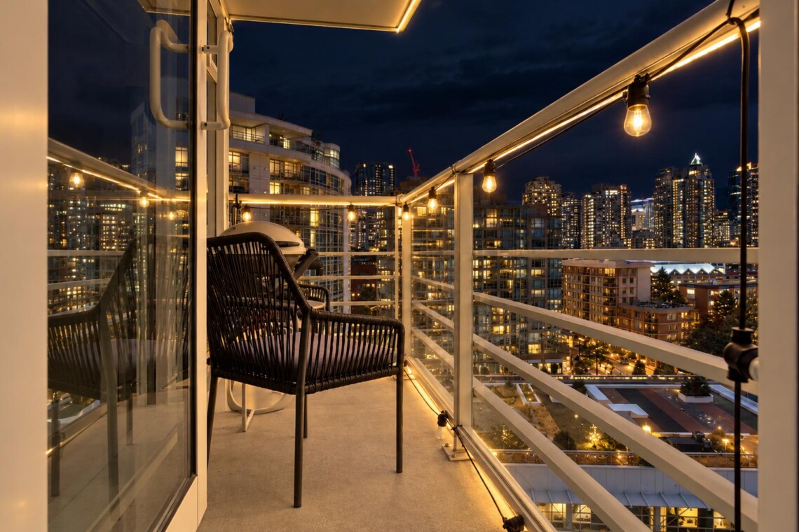 Nighttime balcony with warm string lights, a black wicker chair, and glass railing overlooking a lit city skyline.