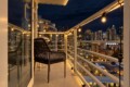 Nighttime balcony with warm string lights, a black wicker chair, and glass railing overlooking a lit city skyline.