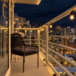 Nighttime balcony with warm string lights, a black wicker chair, and glass railing overlooking a lit city skyline.