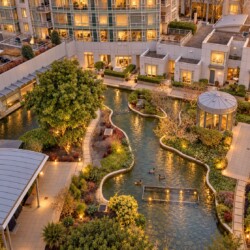 Aerial view of a modern apartment complex with a winding canal, landscaped gardens, and warm indoor lights at dusk, viewed from above.