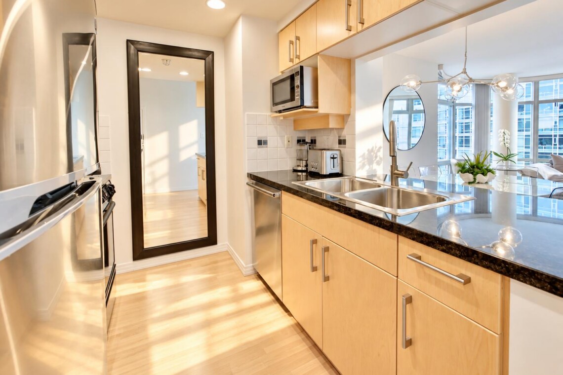 Modern kitchen with light wood cabinets, stainless steel appliances, and a double sink facing a bright living area.