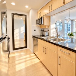 Modern kitchen with light wood cabinets, stainless steel appliances, and a double sink facing a bright living area.