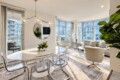 Open-plan living and dining area with a marble dining table, clear acrylic chairs, curved bay windows, and an urban cityscape outside.