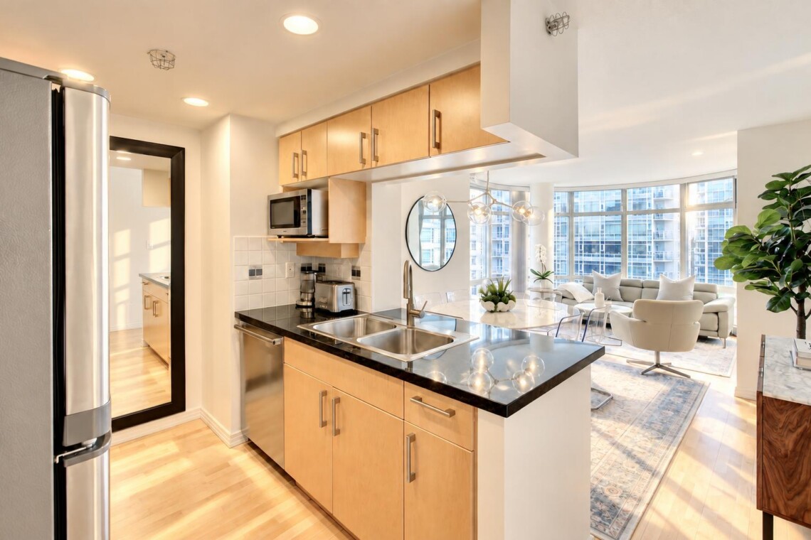 Open-concept kitchen with light wood cabinets, black countertop island, and stainless appliances opening to a bright living room with large windows.
