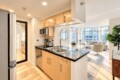 Open-concept kitchen with light wood cabinets, black countertop island, and stainless appliances opening to a bright living room with large windows.