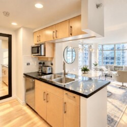 Open-concept kitchen with light wood cabinets, black countertop island, and stainless appliances opening to a bright living room with large windows.