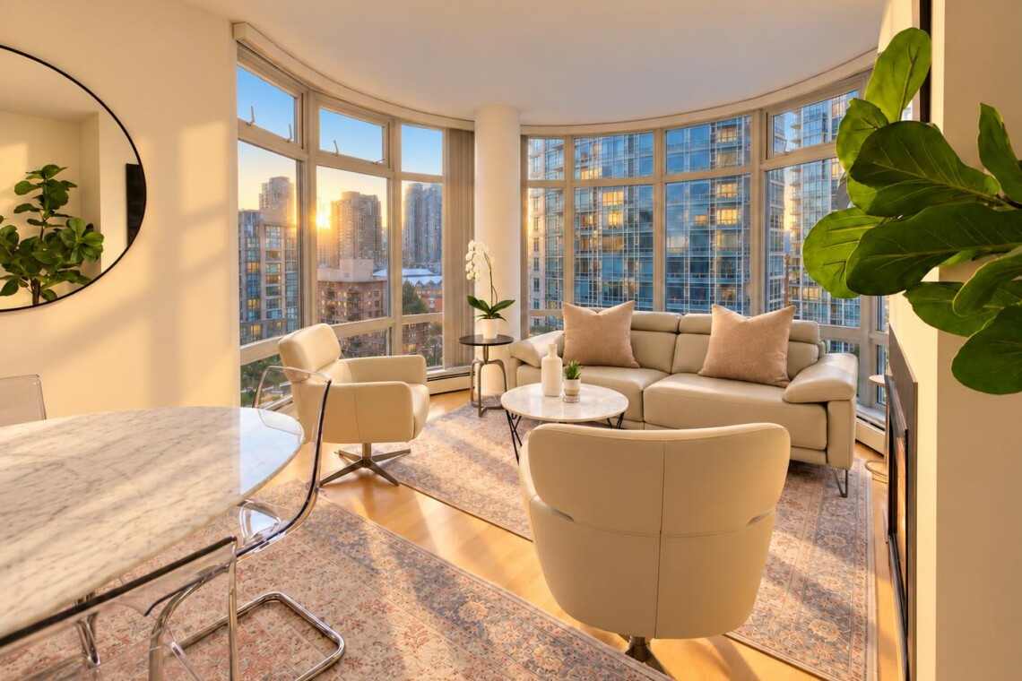 Sunlit modern living room with curved windows and a city skyline view at sunset; beige sofa and chairs, plants, and a marble dining table nearby
