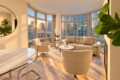 Sunlit modern living room with curved windows and a city skyline view at sunset; beige sofa and chairs, plants, and a marble dining table nearby