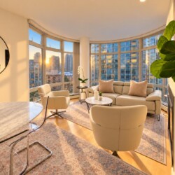Sunlit modern living room with curved windows and a city skyline view at sunset; beige sofa and chairs, plants, and a marble dining table nearby