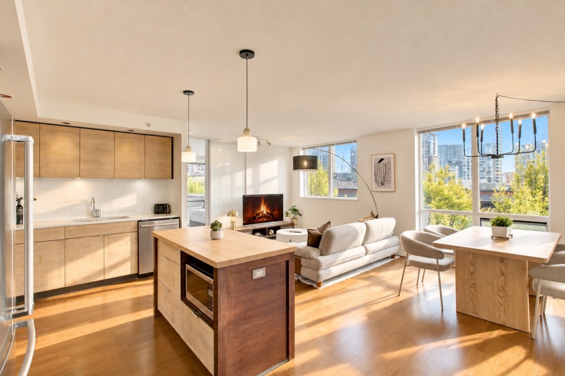Bright open-plan apartment with a wooden kitchen island, wall cabinets, and a living area featuring a sofa, TV, and large windows with a city view on the right side.