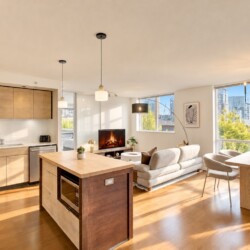 Bright open-plan apartment with a wooden kitchen island, wall cabinets, and a living area featuring a sofa, TV, and large windows with a city view on the right side.