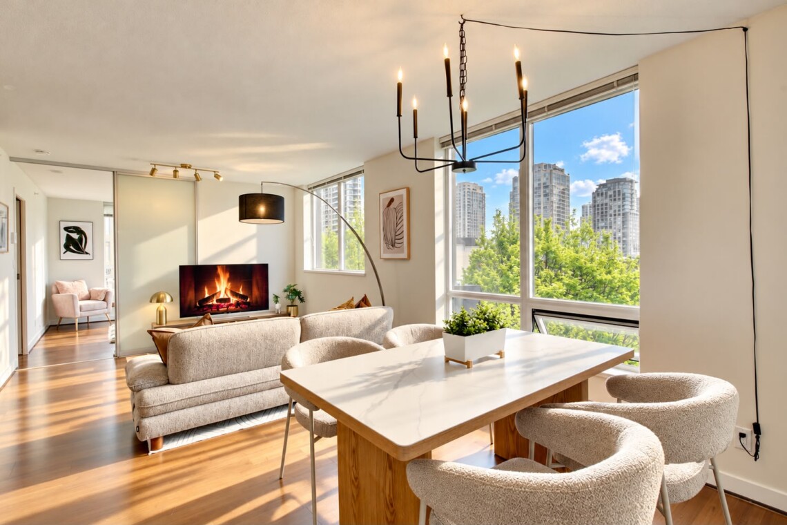 Open-concept living and dining area with a lit fireplace, beige sofa, and large windows overlooking a cityscape and trees.