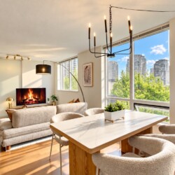 Open-concept living and dining area with a lit fireplace, beige sofa, and large windows overlooking a cityscape and trees.
