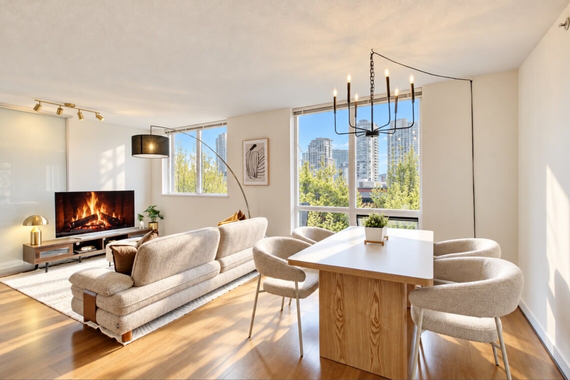 Bright modern living room with a beige sofa, fireplace, TV, and a white dining table by large windows overlooking the city skyline.