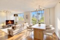 Bright modern living room with a beige sofa, fireplace, TV, and a white dining table by large windows overlooking the city skyline.