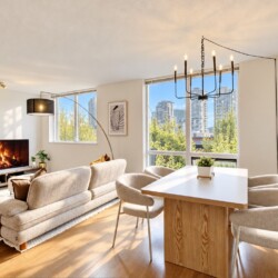 Bright modern living room with a beige sofa, fireplace, TV, and a white dining table by large windows overlooking the city skyline.