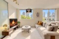 Contemporary living room with a lit fireplace, wooden media console, round coffee table, and beige sectional sofa; large windows show a city skyline.