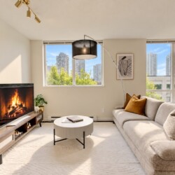 Contemporary living room with a lit fireplace, wooden media console, round coffee table, and beige sectional sofa; large windows show a city skyline.