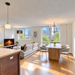 Bright open-concept living area with a fireplace, beige sofa, and large windows overlooking a cityscape and trees.