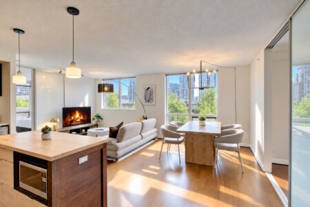 Bright open-concept living area with a fireplace, beige sofa, and large windows overlooking a cityscape and trees.