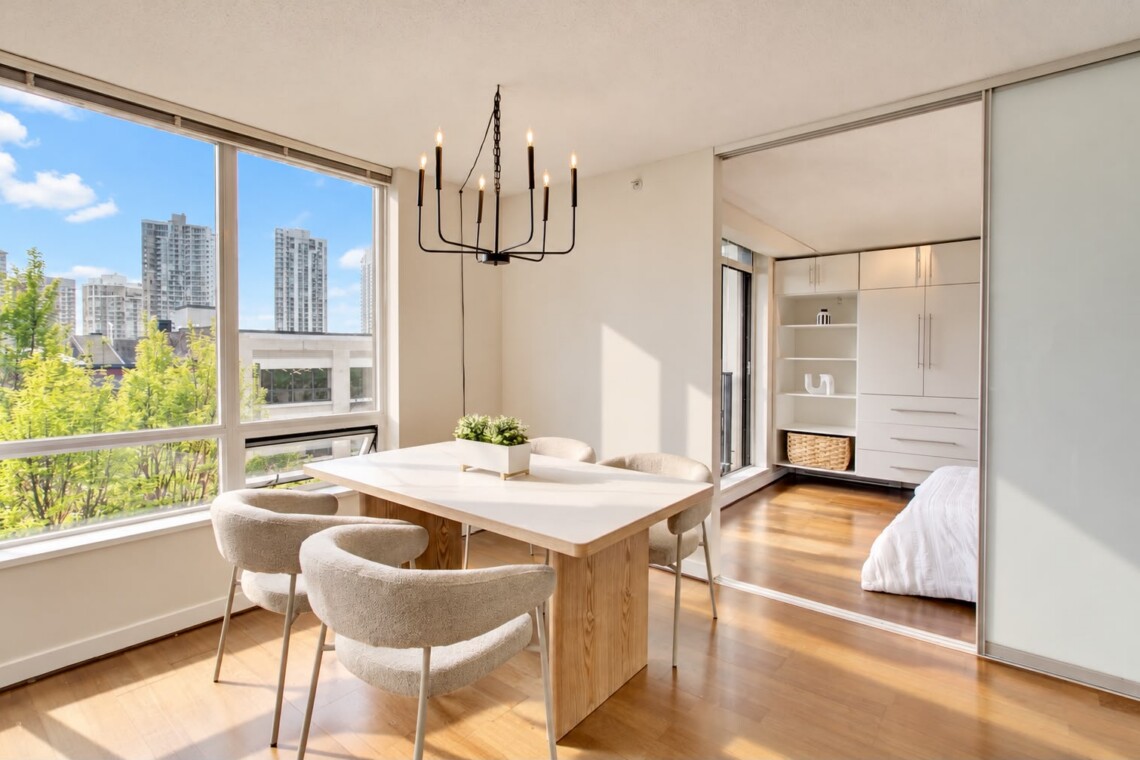 Bright, modern dining area with large windows overlooking a city skyline, a light wood table and cream upholstered chairs, and a black chandelier above.