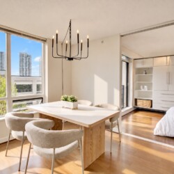 Bright, modern dining area with large windows overlooking a city skyline, a light wood table and cream upholstered chairs, and a black chandelier above.