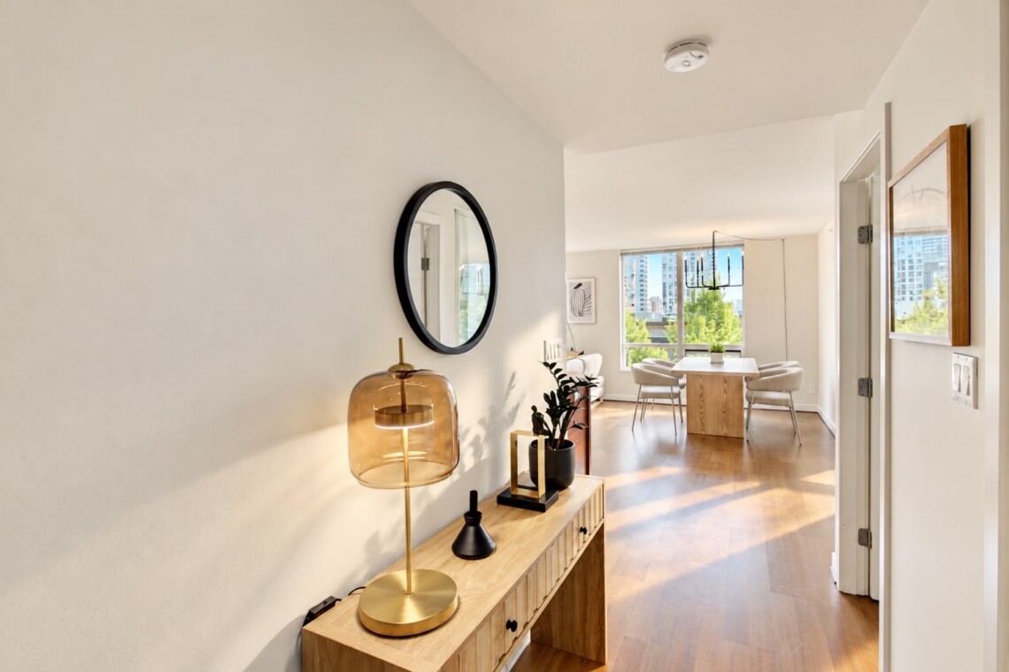 Bright hallway with a wooden console table, amber glass lamp, circular wall mirror, and greenery leading to a dining area by large windows.