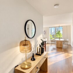 Bright hallway with a wooden console table, amber glass lamp, circular wall mirror, and greenery leading to a dining area by large windows.
