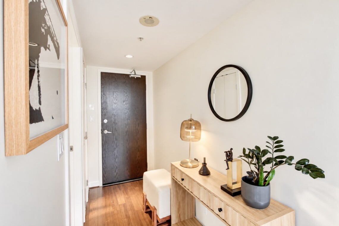 Modern entryway with a wooden console table, round black-framed mirror, amber glass lamp, and a potted plant beside the front door.