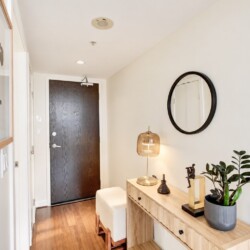 Modern entryway with a wooden console table, round black-framed mirror, amber glass lamp, and a potted plant beside the front door.