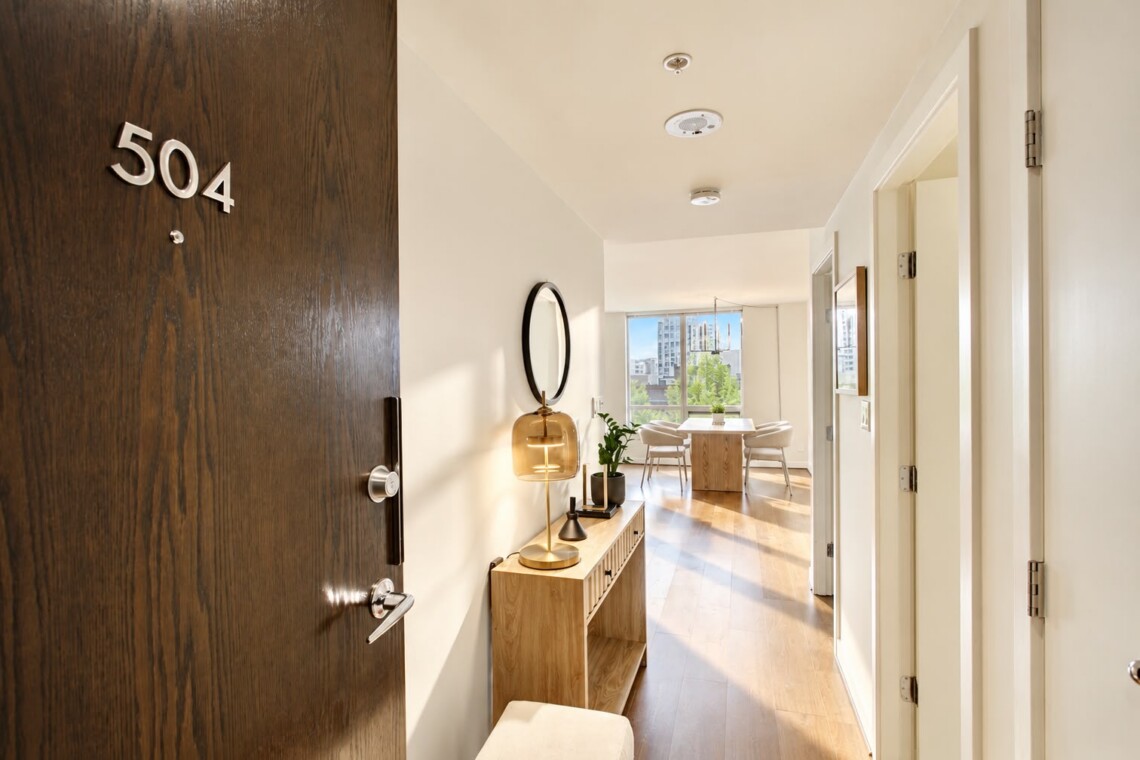 Apartment entry hallway with a dark wood door marked 504, a wooden console, round mirror, and warm lamp.