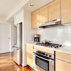 Modern kitchen with stainless steel refrigerator, gas range, and light wood cabinets, white tile backsplash, and wooden cutting boards on the counter