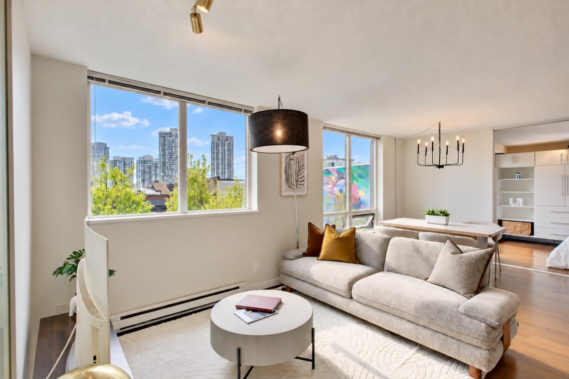 Bright open-plan living room with a beige sectional, mustard cushions, round coffee table, and large windows showing the city skyline.
