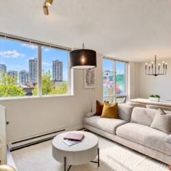 Bright open-plan living room with a beige sectional, mustard cushions, round coffee table, and large windows showing the city skyline.